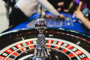 Vibrant casino table with roulette in motion, with casino chips, tokens, the hand of croupier, dollar money and a group of gambling rich people playing bet, blue poker table and deck of cards